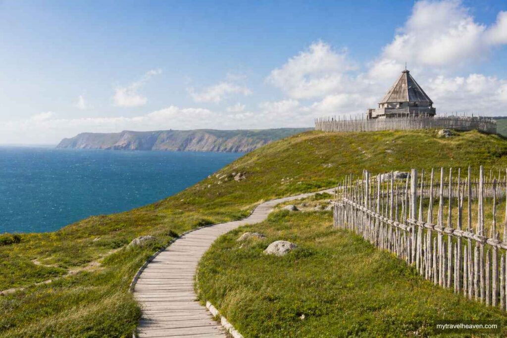 Wooden boardwalk along the Viking Trail in Newfoundland and Labrador with grassy hills, coastal cliffs, and ocean views under a blue sky.