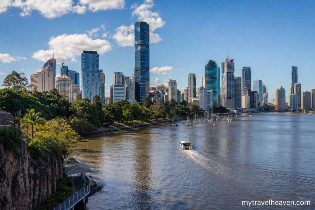 Panoramic riverside view of Brisbane skyline along the Brisbane River, featuring modern skyscrapers, greenery, and a ferry cruising on a sunny day.