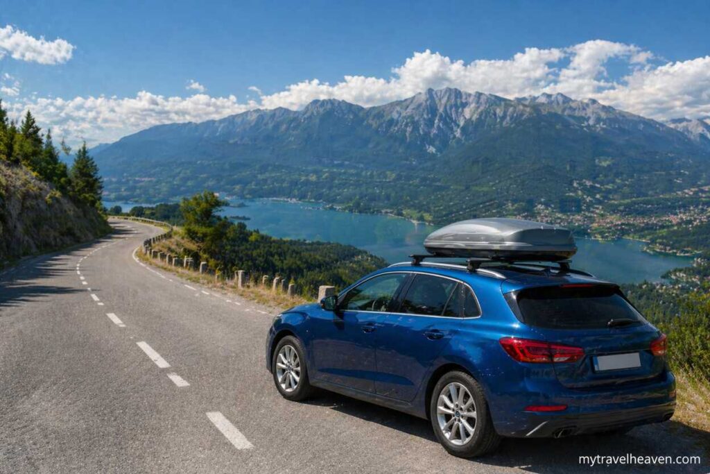 Blue car with a rooftop cargo box parked on a scenic European mountain road overlooking a lake and mountains, with mytravelheaven.com watermark in the bottom right corner.