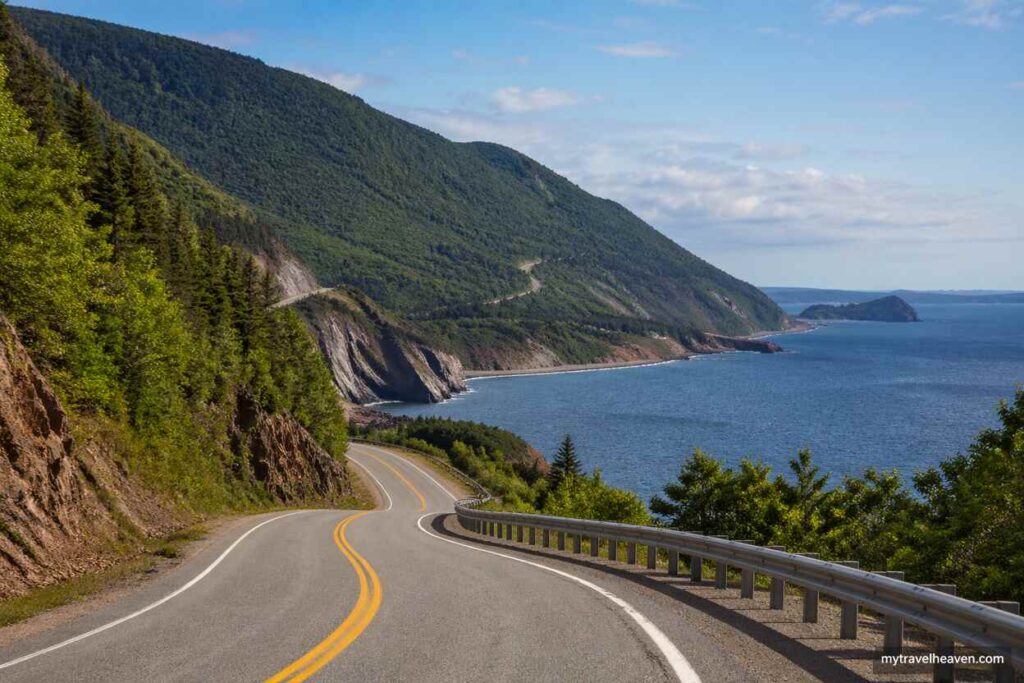 Scenic coastal drive along the Cabot Trail in Nova Scotia with a winding road, green mountains, and blue ocean under a clear sky.