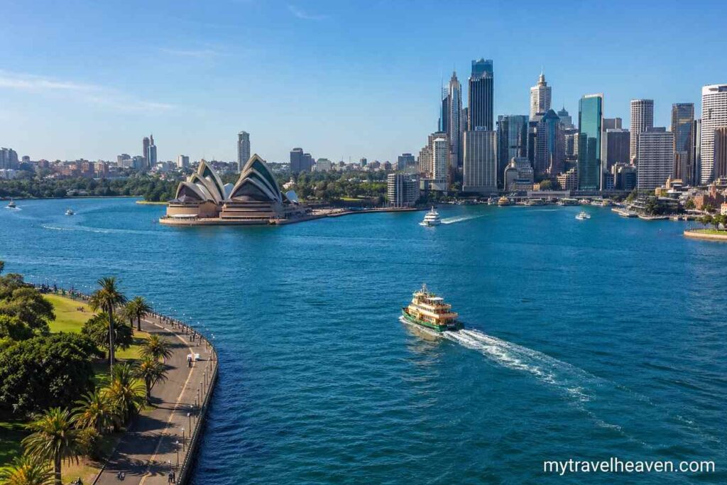 Panoramic view of Sydney harbor featuring the Sydney Opera House, city skyline, blue waters, and a ferry boat on a sunny day.