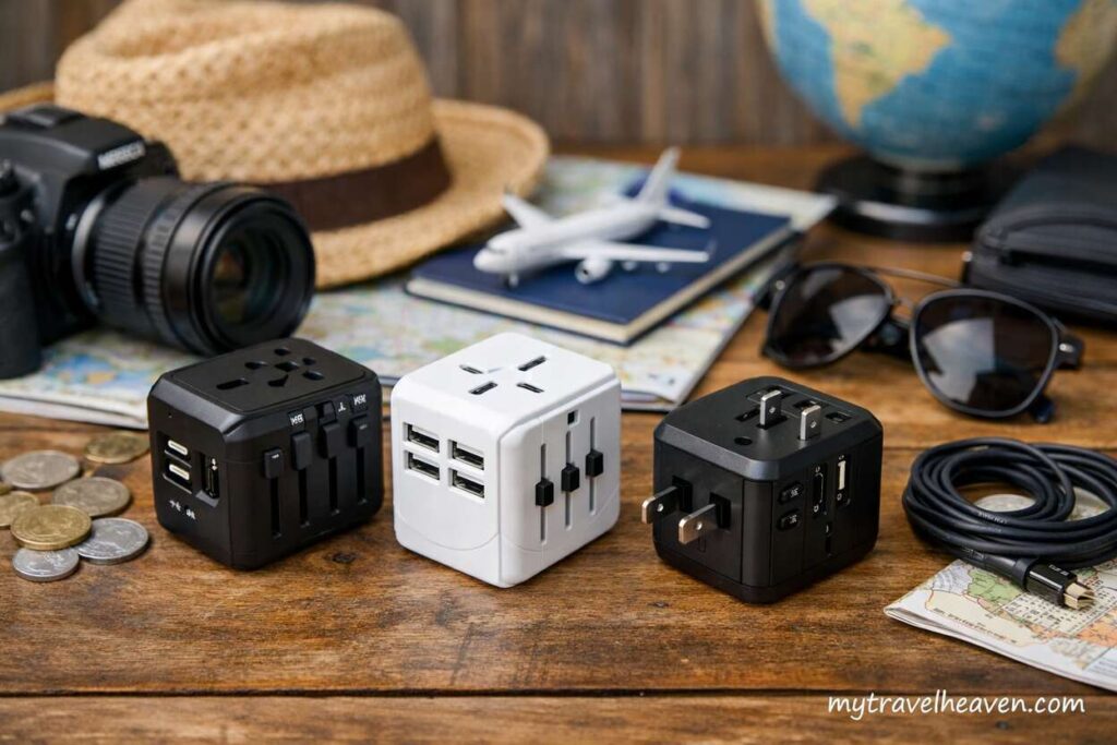 Universal travel adapters with multiple plug types placed on a wooden table alongside travel essentials like a camera, sunglasses, passport, and world map, with “mytravelheaven.com” visible in the corner.