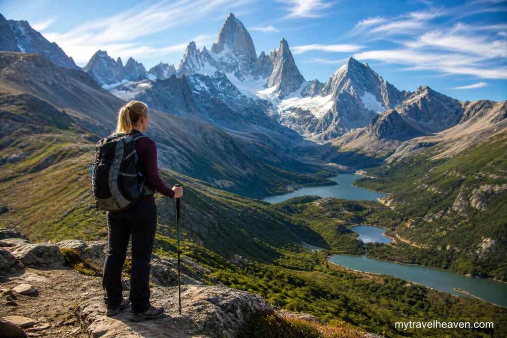 Hiker standing on a mountain viewpoint overlooking a scenic valley with lakes and snow-capped peaks under a clear blue sky.