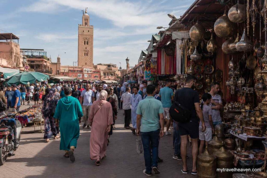 Bustling Marrakech market in Morocco with crowded walkways, colorful stalls, traditional lanterns, and the Koutoubia Mosque visible in the background.