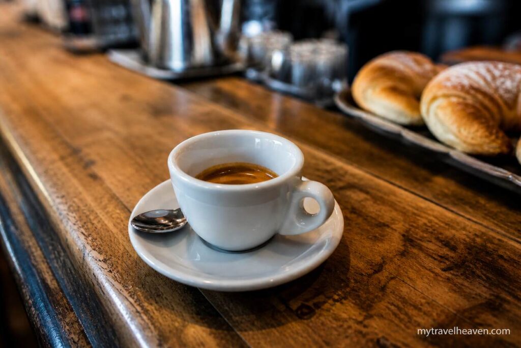 Italian espresso served in a white cup on a rustic wooden café counter with croissants in the background.