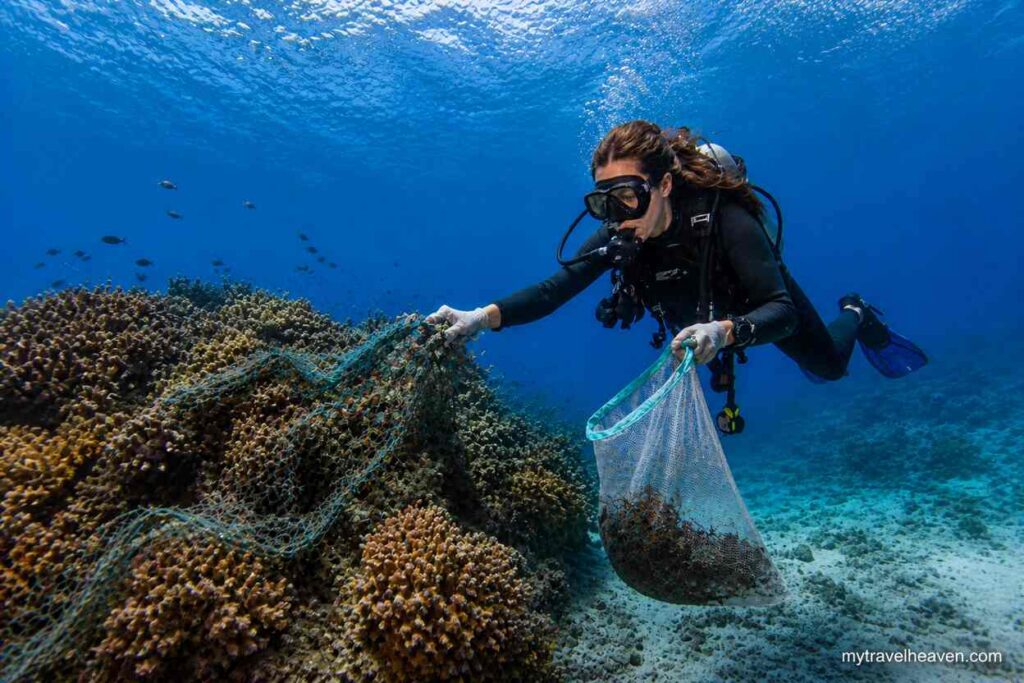 Scuba diver removing fishing net debris from a coral reef while collecting waste in a mesh bag underwater.