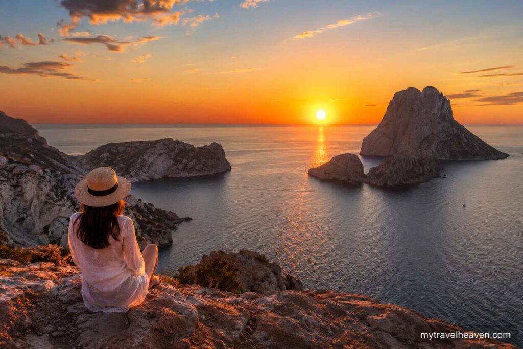 Golden sunset over a rocky coastal landscape with a traveler sitting on a cliff overlooking calm ocean waters and scenic islands during sunset.