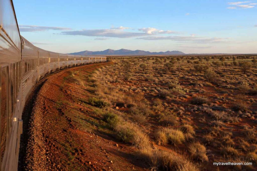 A long train crossing the Australian Outback through red desert terrain, scattered shrubs, and distant rocky mountains under a warm evening sky.