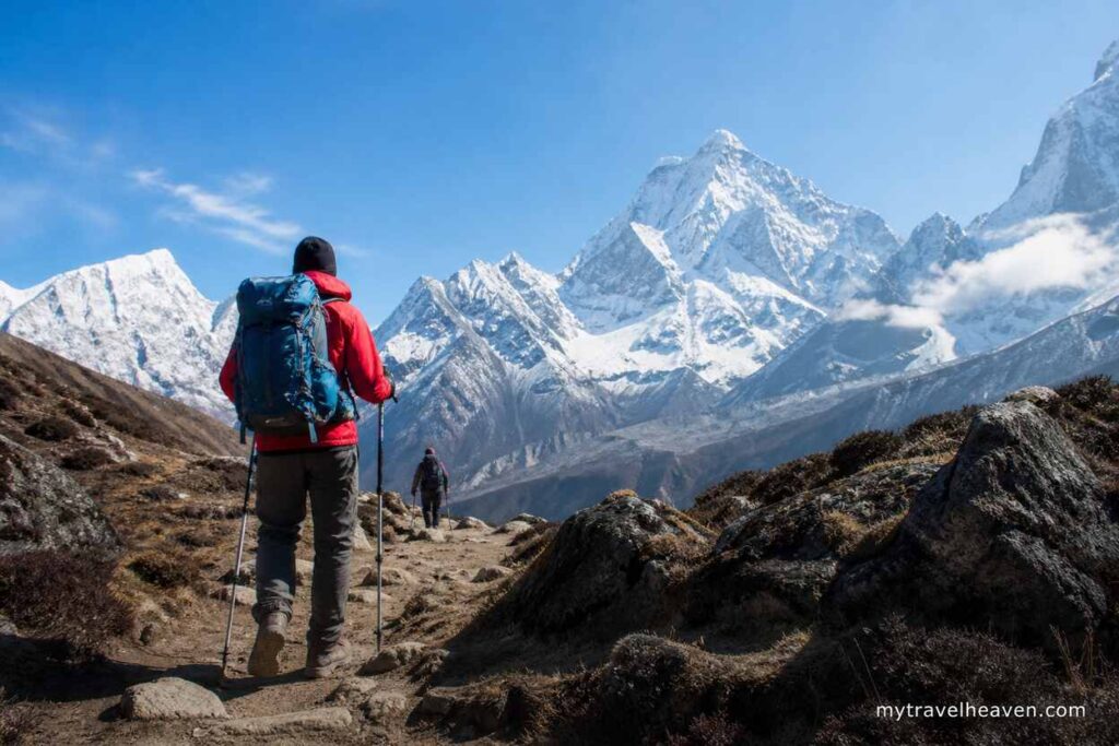 Hikers trekking on a rocky mountain trail with snow-covered peaks in the background under a clear blue sky, symbolizing adventure travel and trekking insurance coverage.