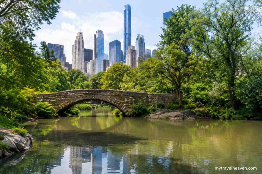 Visitors enjoying a scenic view of Central Park with a stone bridge, calm lake, lush green trees, and the New York City skyline in the background.
