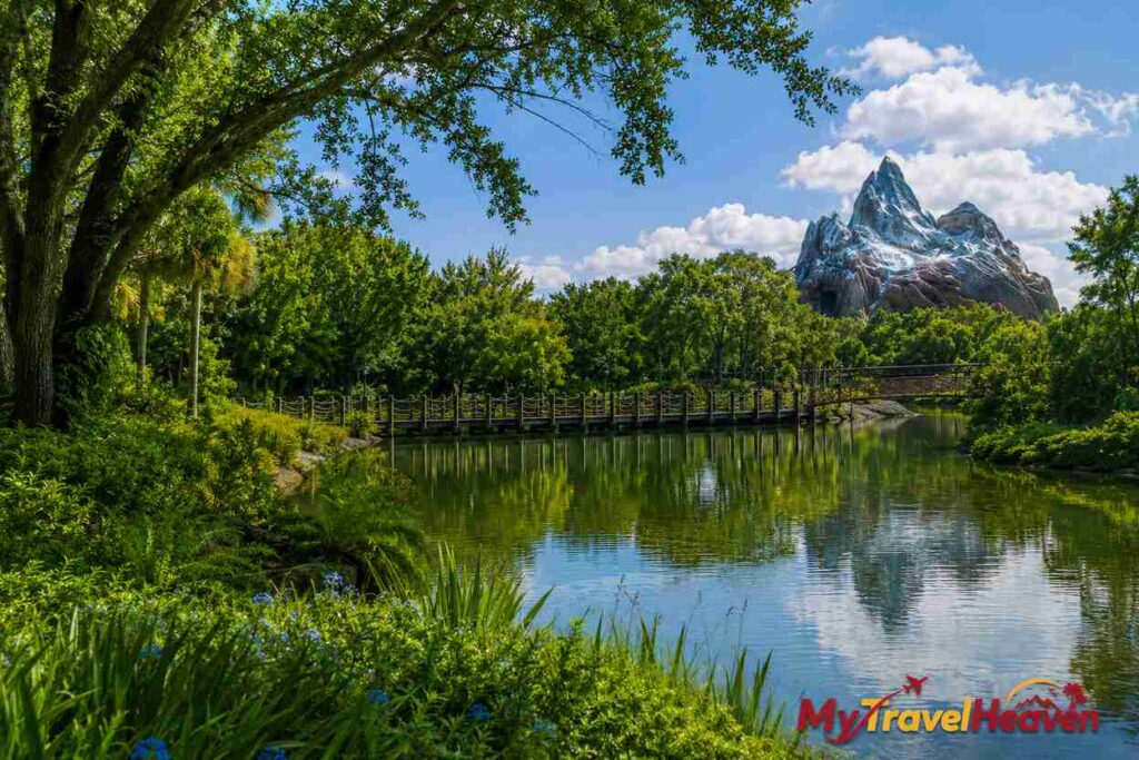 Lush natural landscape inside Disney World featuring a peaceful lake, wooden bridge, dense greenery, and mountain backdrop with the MyTravelHeaven logo in the bottom-right corner.